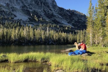 This photo shows a backpacker sitting by a mountain lake drinking coffee with the Sierra Designs Backcountry Quilt 700.