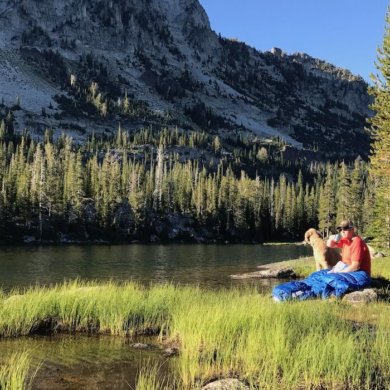 This photo shows a backpacker sitting by a mountain lake drinking coffee with the Sierra Designs Backcountry Quilt 700.