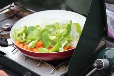 This photo shows a camp stove with a frying pan full of veggies frying outside.