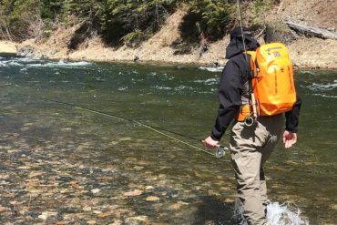 This image shows a fly fisher wearing a Fishpond Thunderhead Submersible Backpack with a Quickshot Rod Holder accessory while wading in a river.