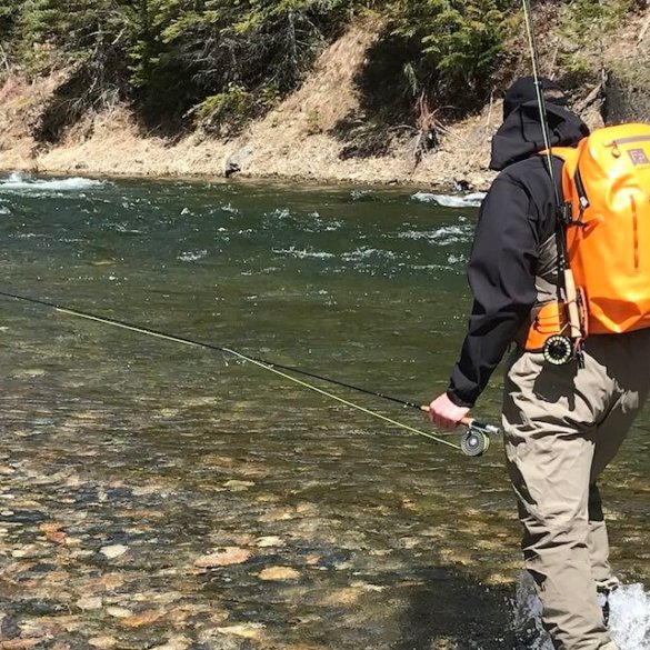 This image shows a fly fisher wearing a Fishpond Thunderhead Submersible Backpack with a Quickshot Rod Holder accessory while wading in a river.