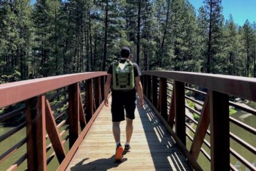 This photo shows the ICEMULE Pro cooler being worn by a hiker on a bridge outside near trees and a lake.