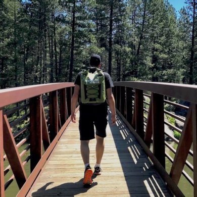 This photo shows the ICEMULE Pro cooler being worn by a hiker on a bridge outside near trees and a lake.
