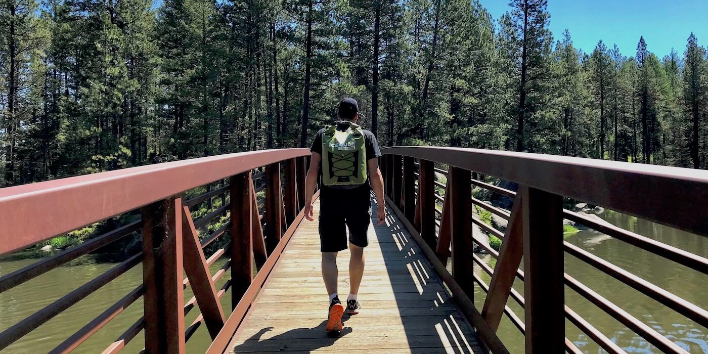 This photo shows the ICEMULE Pro cooler being worn by a hiker on a bridge outside near trees and a lake.