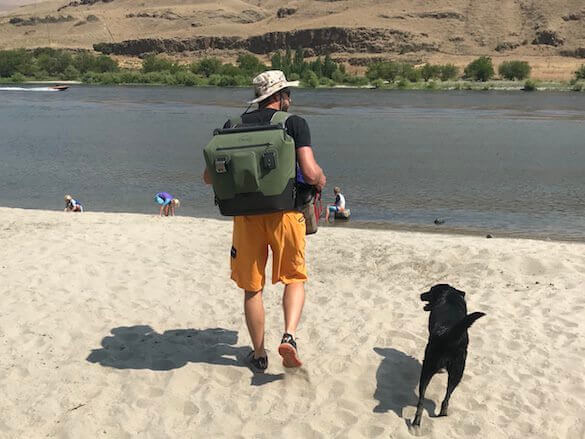 This photo shows a man carrying the OtterBox Trooper LT 30 backpack cooler on a beach near a river.