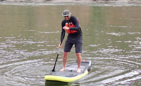 This Shelta Hat review photo shows a man wearing the Shelta Raptor V2 hat while paddle boarding on a river.