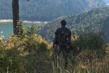 This best bowhunting gift photo shows a bowhunter standing with a hunting bow on a ridge overlooking a lake.