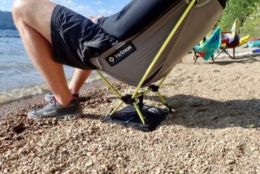 This photo shows a man sitting in the Helinox Chair Zero with Ground Sheet on a beach by a lake.