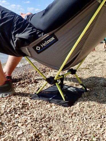 This photo shows a man sitting in the Helinox Chair Zero with Ground Sheet on a beach by a lake.