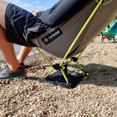 This photo shows a man sitting in the Helinox Chair Zero with Ground Sheet on a beach by a lake.