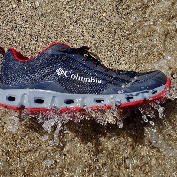 This testing and review water shoe photo shows a closeup of a Columbia Drainmaker IV water shoe on a beach getting hit by a wave.
