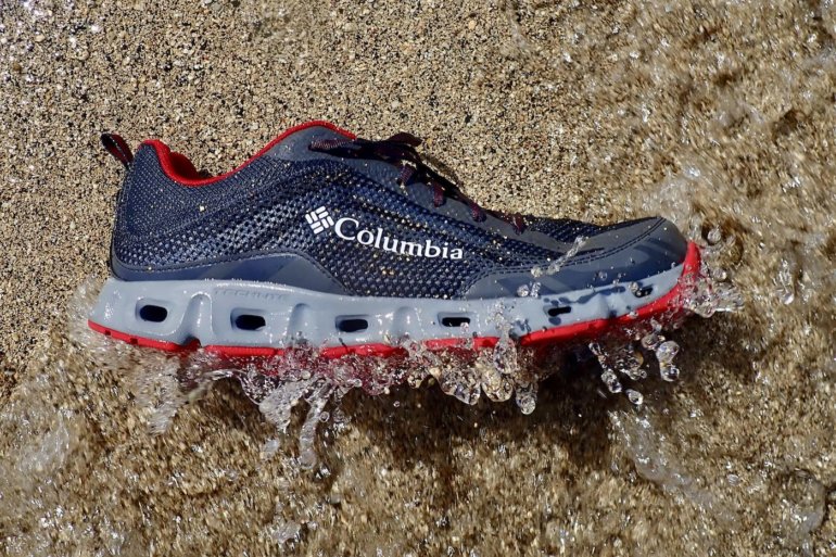 This testing and review water shoe photo shows a closeup of a Columbia Drainmaker IV water shoe on a beach getting hit by a wave.