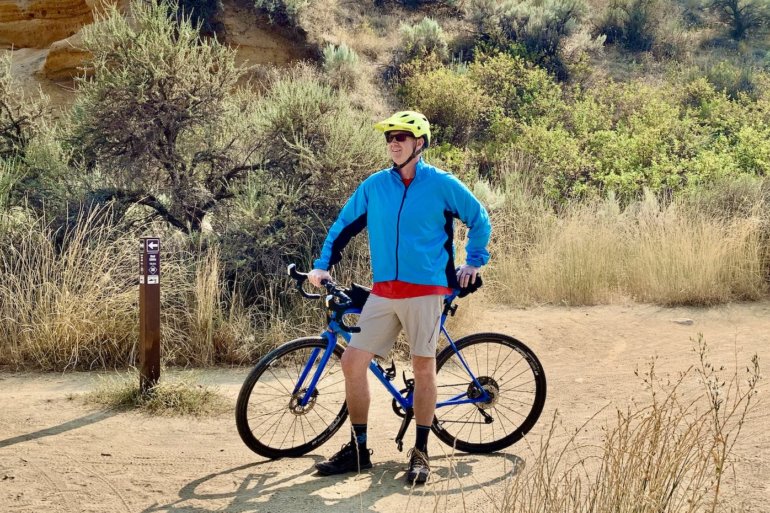 This review and testing photo shows the author wearing the Showers Pass Ultralight Wind Jacket on a trail with a gravel bike.