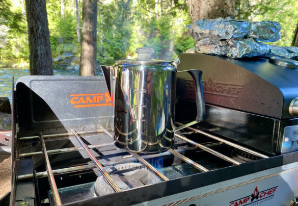 This camping photo shows a camping stove with a coffee pot outside in a forest.