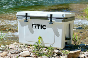This camping gift photo shows the RTIC Ultra-Light Cooler outside near a stream at a campsite.