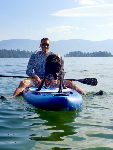 This outdoor gift guide photo shows the author outside on a lake on a paddle board with a dog on a summer day.
