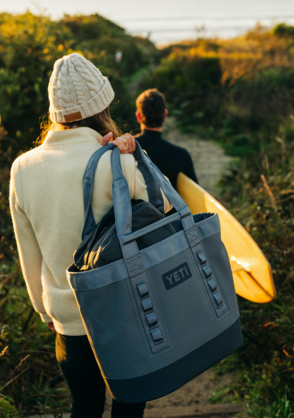 This outdoor gear photo shows the YETI Camino 35 Carryall tote bag being carried by a woman heading down a trail to a beach.