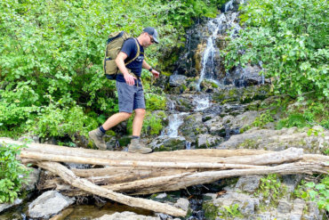 This photo shows the author wearing KUHL Renegade Shorts while crossing a creek on a log outside while hiking during the testing and review process.