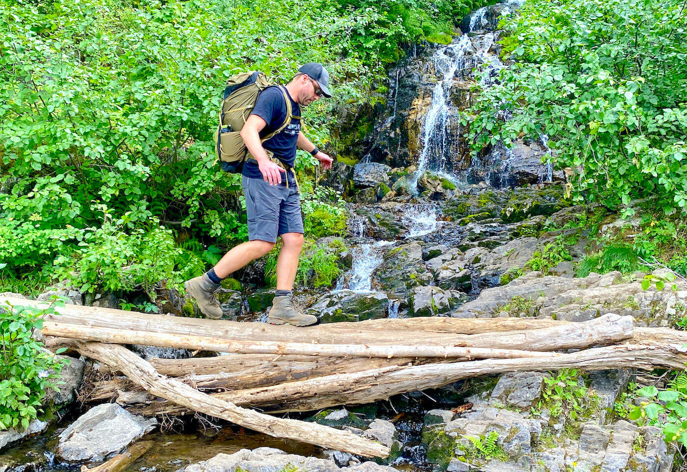 This photo shows the author wearing KUHL Renegade Shorts while crossing a creek on a log outside while hiking during the testing and review process.