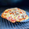 This photo shows a homemade pizza being lifted from a Traeger pellet grill with a pizza paddle.