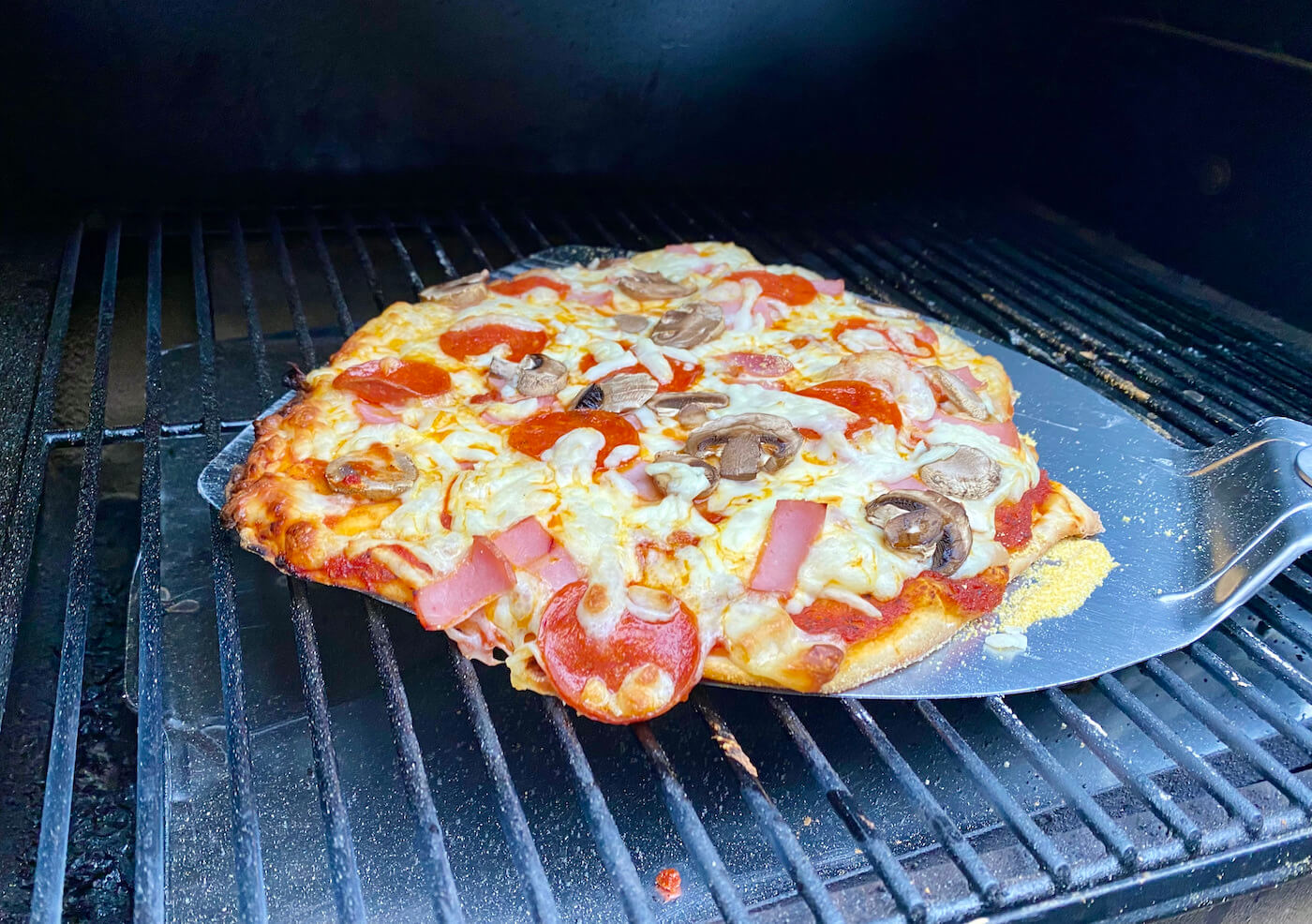 This photo shows a homemade pizza being lifted from a Traeger pellet grill with a pizza paddle.
