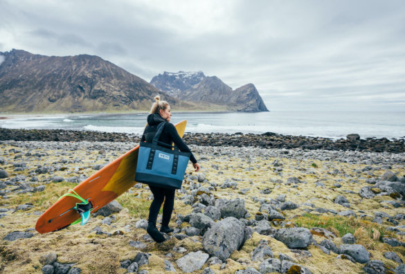 This photo shows a woman near a northern sea carrying a Nordic Blue YETI Camino Carryall tote bag with a surf board.