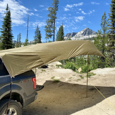 This photo shows the SJK Roadhouse Tarp set up on the back of a pickup outside in a forest near mountains.