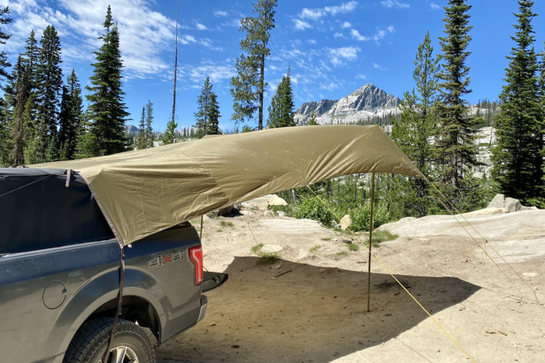 This photo shows the SJK Roadhouse Tarp set up on the back of a pickup outside in a forest near mountains.