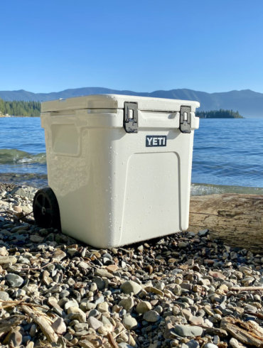 This photo shows the YETI Roadie 48 Wheeled Cooler on a beach near a lake.