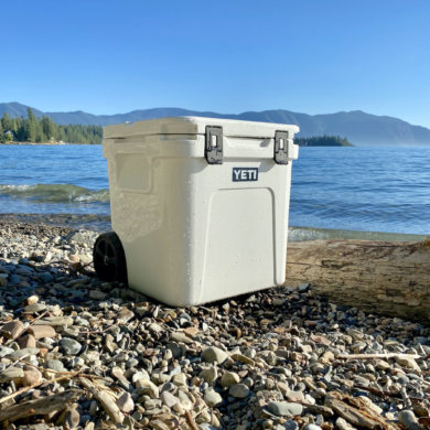 This photo shows the YETI Roadie 48 Wheeled Cooler on a beach near a lake.