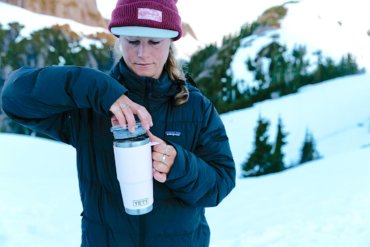 This photo shows a woman opening an Ice Pink YETI Rambler Tumbler outside in a winter landscape.