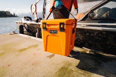 This photo shows a man carrying a YETI Roadie 24 Hard Cooler on a dock near a boat.