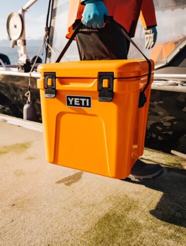 This photo shows a man carrying a YETI Roadie 24 Hard Cooler on a dock near a boat.