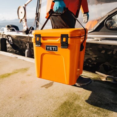 This photo shows a man carrying a YETI Roadie 24 Hard Cooler on a dock near a boat.