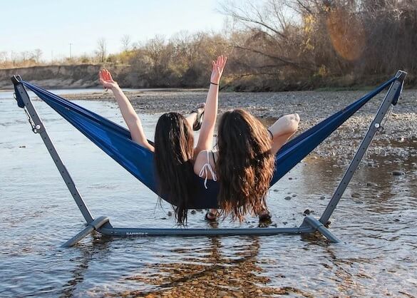 This photo shows the Kammok Swiftlet Hammock Stand with two women sitting in a hammock outside near a river.