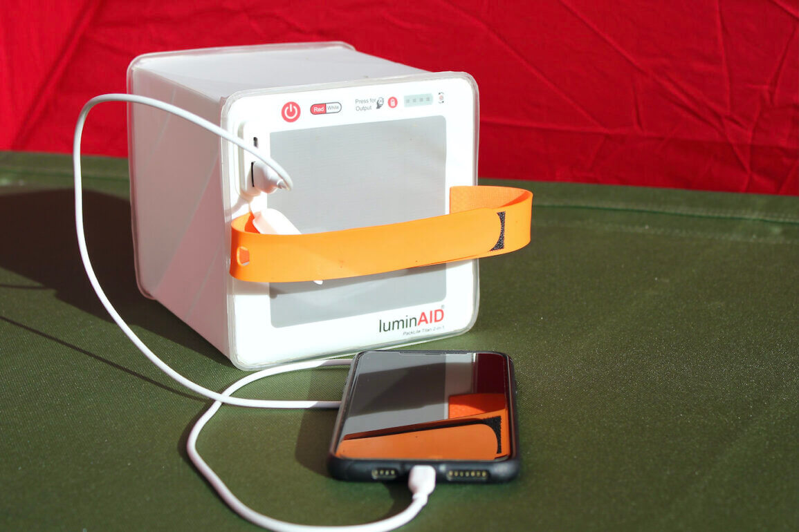 This photo shows the LuminAid PackLite Titan 2-in-1 Solar Lantern Charger charging a mobile phone on a camping cot in a tent.