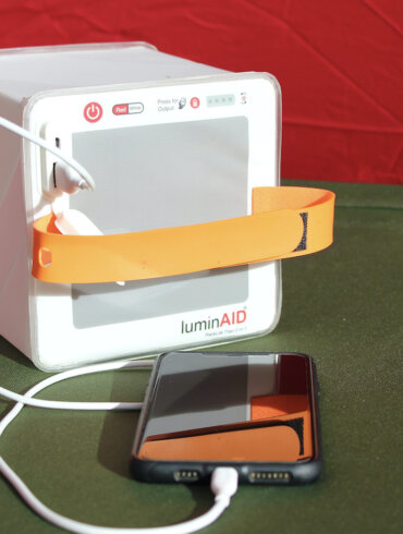 This photo shows the LuminAid PackLite Titan 2-in-1 Solar Lantern Charger charging a mobile phone on a camping cot in a tent.