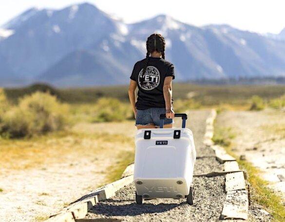 This photo shows an outdoorsy woman pulling a YETI Roadie 48 Wheeled Cooler down a gravel path toward mountains.