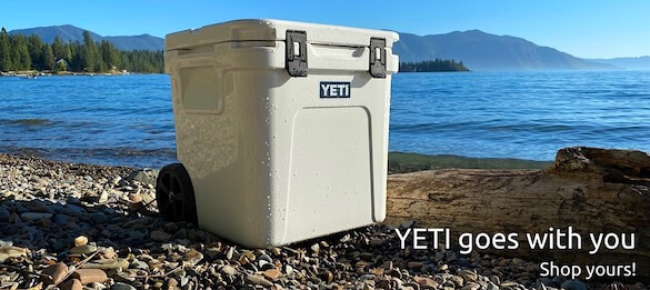 This image shows a YETI Roadie 48 wheeled cooler on a beach with a lake in the background.