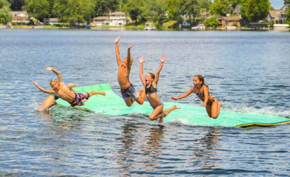This photo shows children jumping off of an Aqua Lily Bullfrog Lily Pad on a lake.