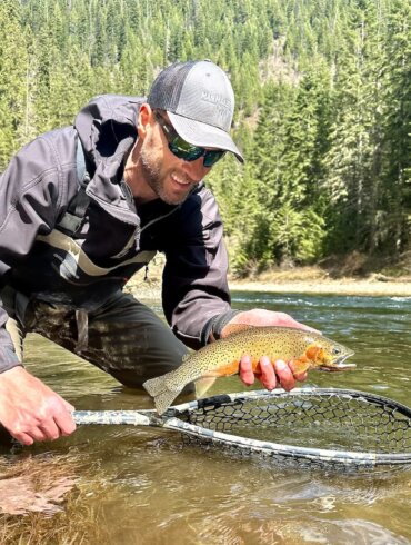 The author wearing the Smith Guide's Choice sunglasses while holding a cutthroat trout over a fly fishing net in a river.