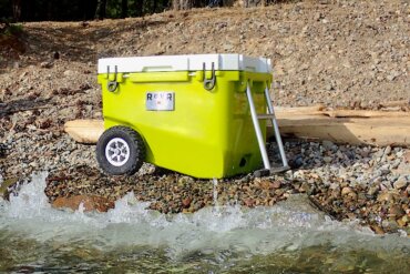 This review photo shows the RovR RollR 60 Wheeled Cooler on a rocky beach near a lake during the testing and review process.