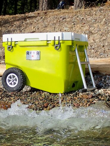 This review photo shows the RovR RollR 60 Wheeled Cooler on a rocky beach near a lake during the testing and review process.