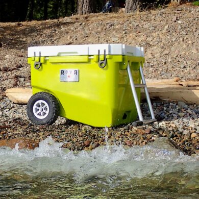 This review photo shows the RovR RollR 60 Wheeled Cooler on a rocky beach near a lake during the testing and review process.