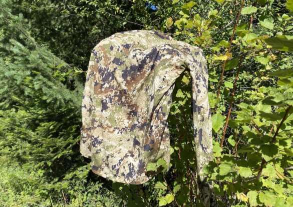 This photo shows a Sitka Core Merino 120 LS Crew shirt drying on a tree branch in the sun in a forest.