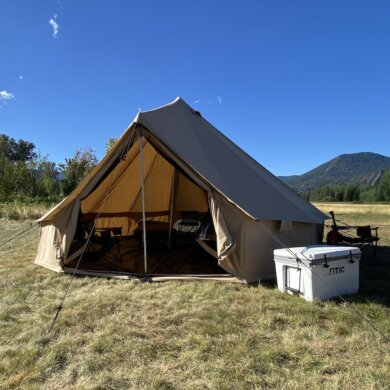 This photo shows a camping tent with camp chairs and cots inside near a camping table.