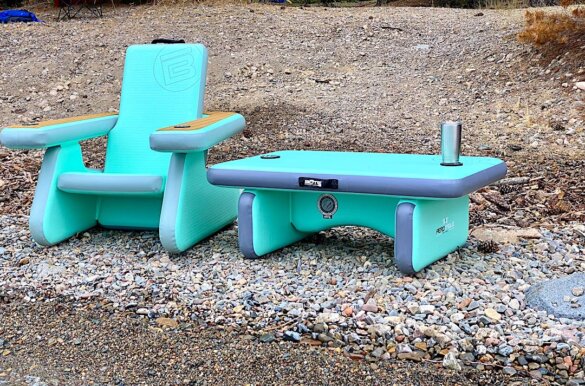This photo shows the BOTE Inflatable Aero Table next to an Inflatable AeroRondak Chair on a beach near a mountain lake.