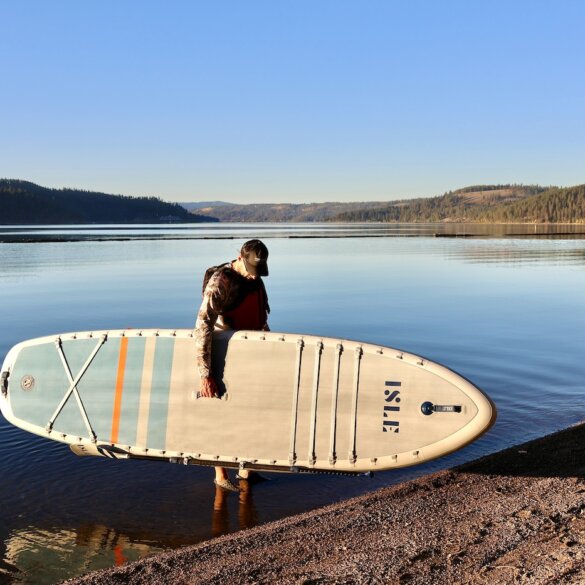 This photo shows the author holding the ISLE Pioneer Pro inflatable standup paddle board in the water next to a beach during the testing and review process.