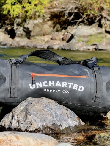 This review photo shows the Uncharted Supply Co The Vault 65L Duffel bag on a rock near a river taken during the author's testing and review process.