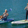 Man and woman each paddling on a new ISLE Pioneer 3 inflatable standup paddle board on a lake.